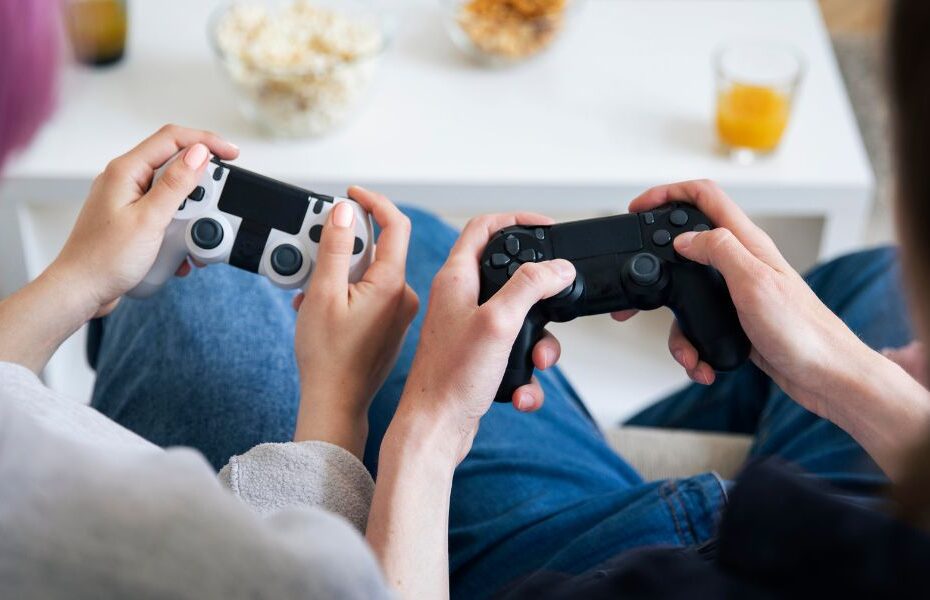 Duas pessoas de costas para a câmera, sentadas em um sofá, segurando controles de videogame. A frente deles há uma mesa de centro com copos de suco e tigelas com pipoca e salgadinho.