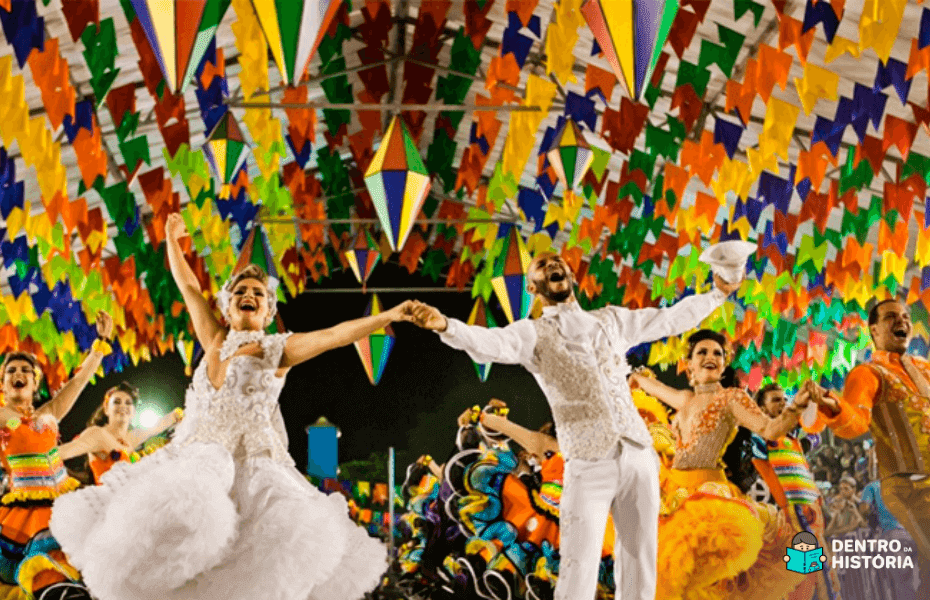 Mulher e homem vestidos de branco dançando em um ambiente decorado por bandeirinhas coloridas.