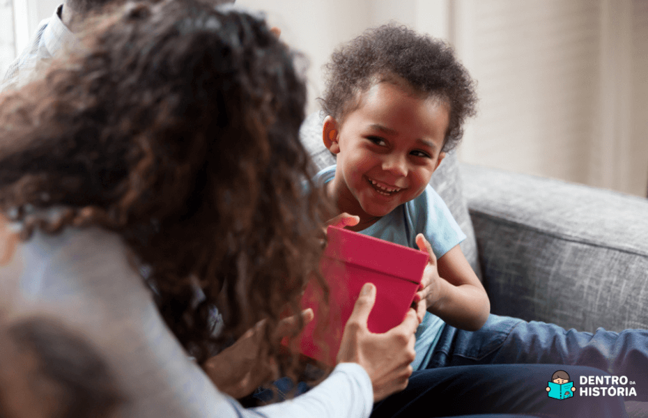 Criança negra sorrindo enquanto recebe uma caixa de presente dos pais.